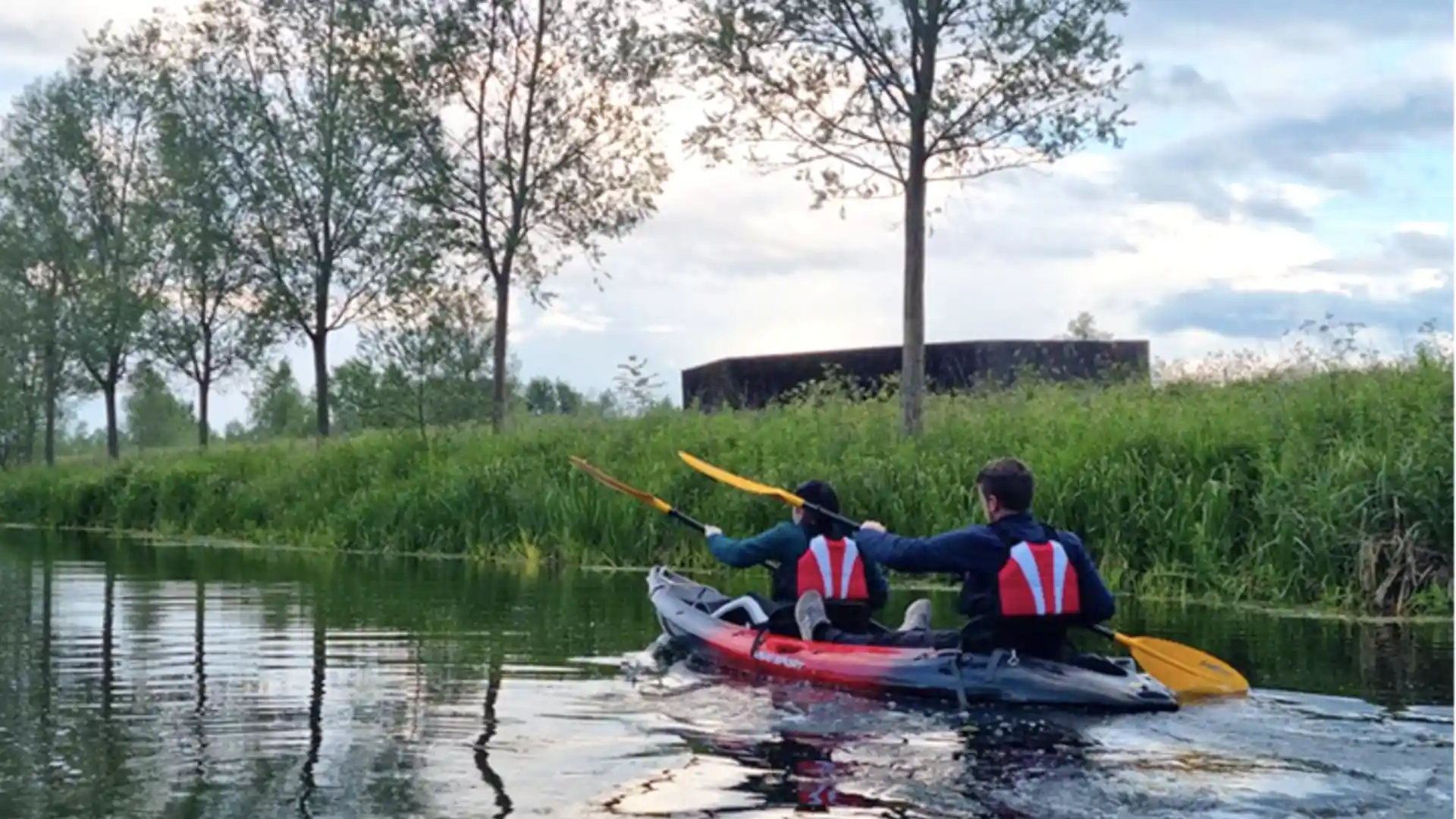 Two people kayaking on the River Stour surrounded by tall grasses and trees in the Dedham Vale National Landscape, with calm reflections on the water at sunset.