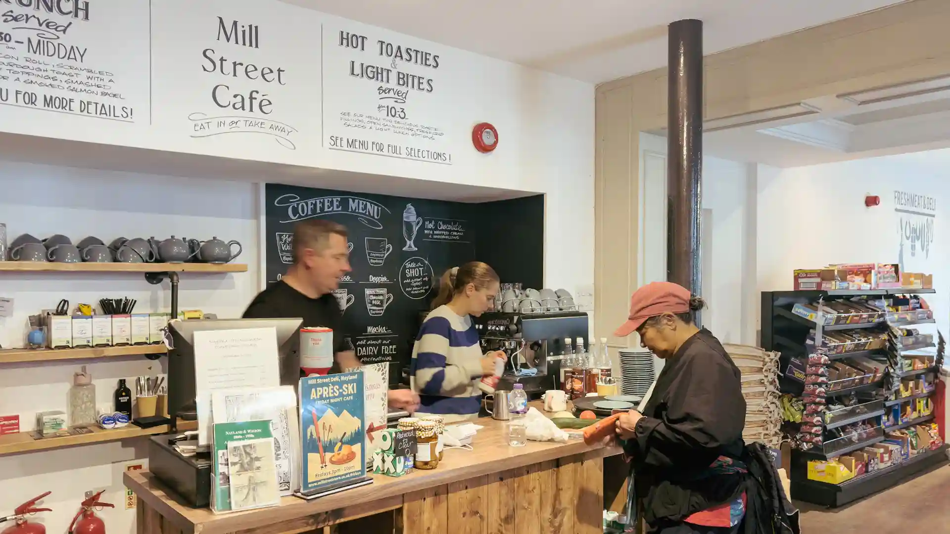 Staff prepare drinks behind the counter at Mill Street Café, with shelves of local produce and handwritten menu boards creating a warm, village café atmosphere.