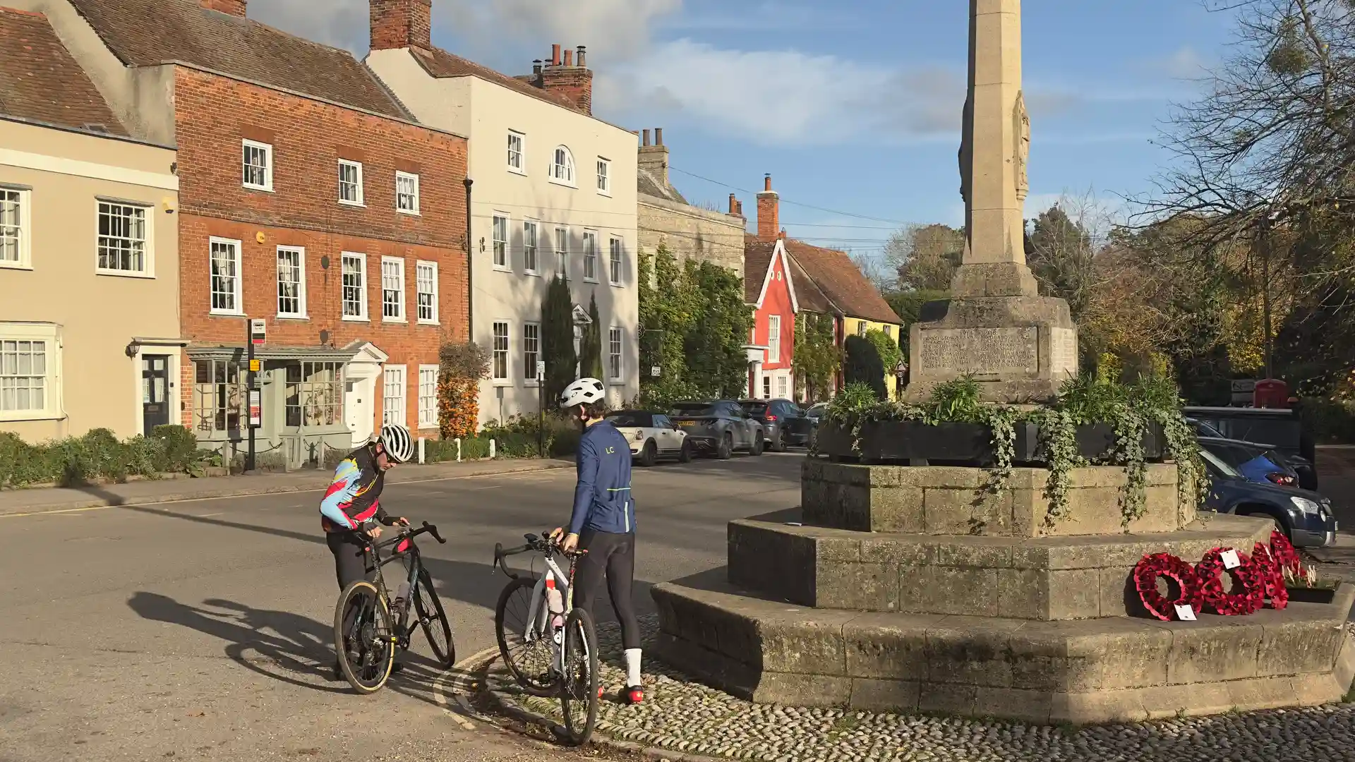 Two cyclists pause with their bikes beside a village war memorial in a historic Suffolk square, with colourful cottages and autumn light in the background.