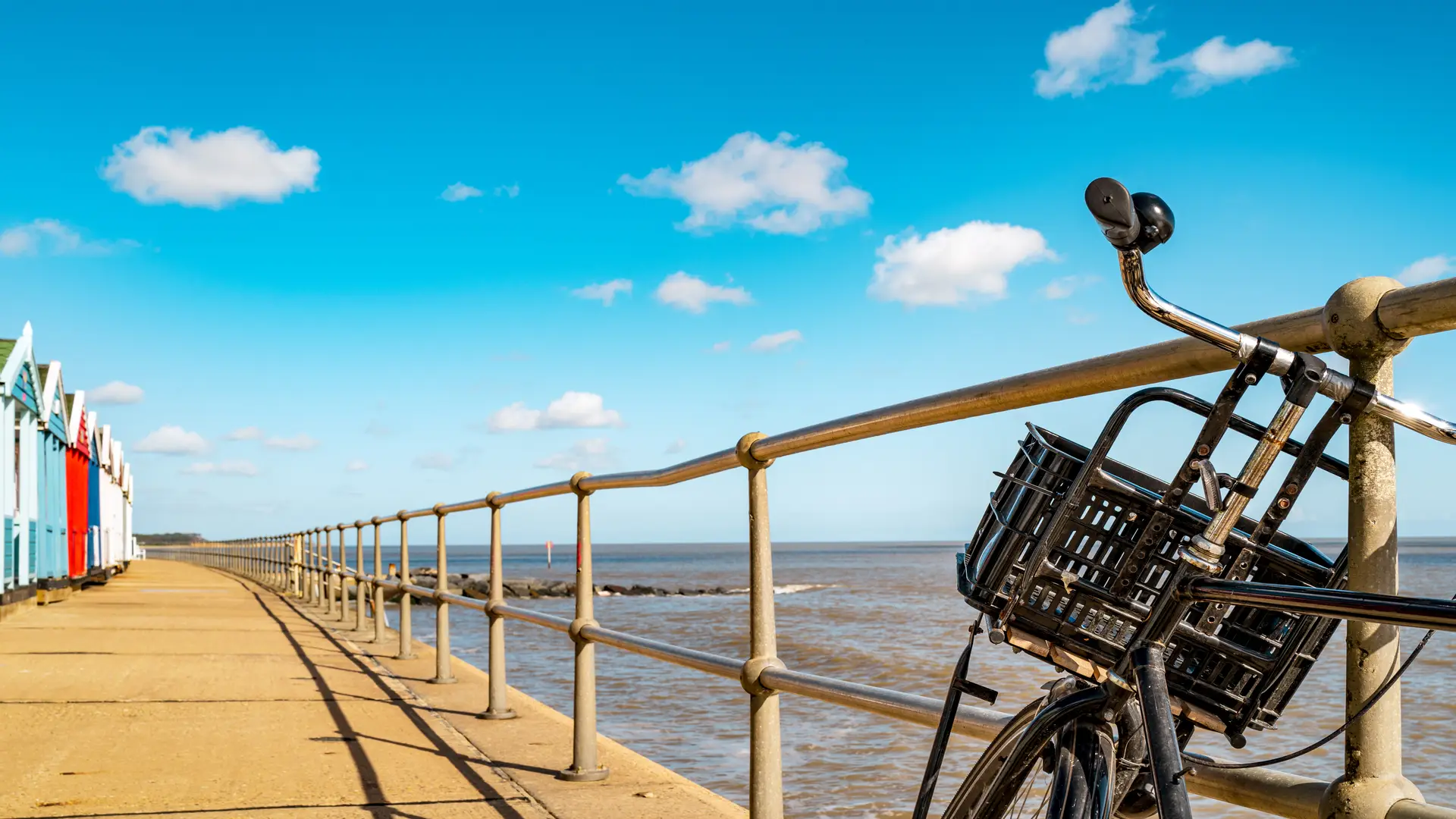 A bicycle resting on a seaside promenade beside colourful beach huts on the Suffolk coast.