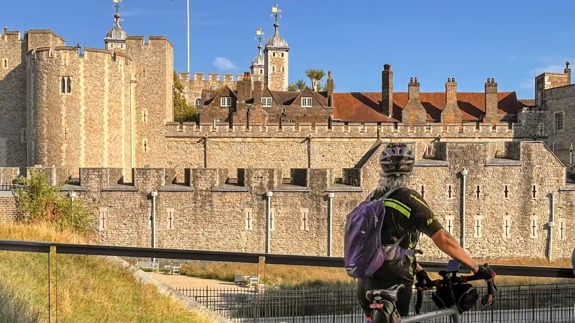 A cyclist pauses beside the Thames Path, looking toward the medieval stone walls and towers of the Tower of London on a bright, clear day.