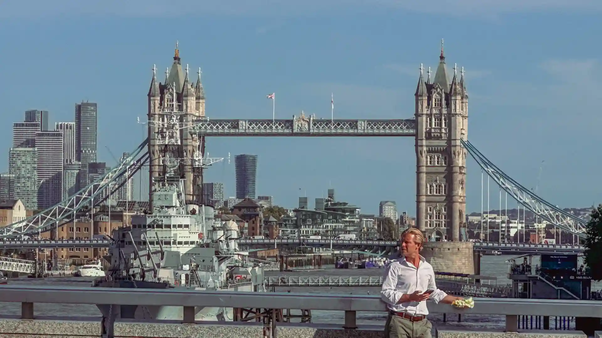 Man standing beside the Thames with Tower Bridge and HMS Belfast in the background on a clear day.