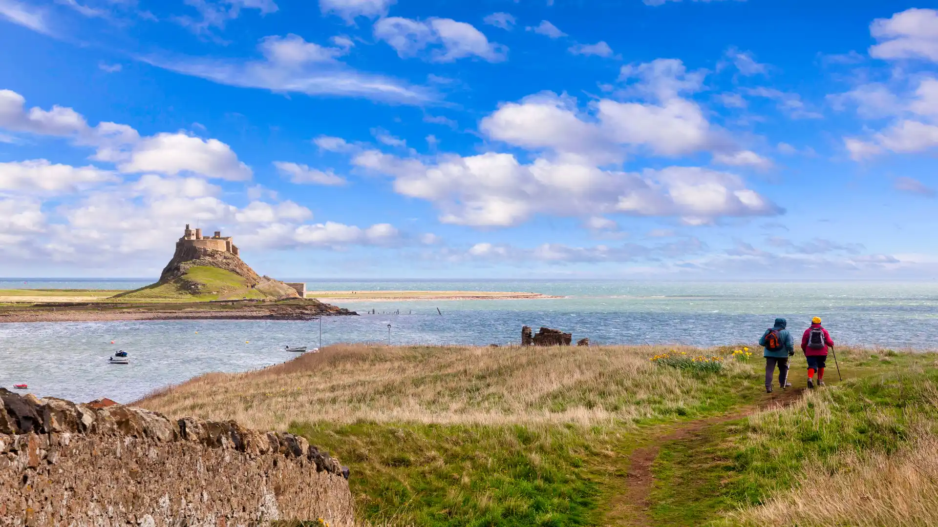 Walkers on a coastal path approaching Lindisfarne Castle on Holy Island, with blue skies, grassy dunes, and the Northumberland Coast National Landscape.