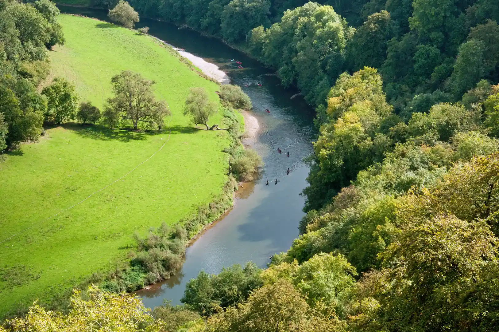 Canoeists paddling along a winding stretch of the River Wye, surrounded by green meadows and dense woodland in the Wye Valley National Landscape.