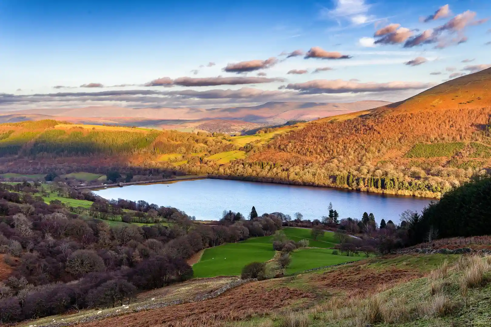 A wide landscape view of the Brecon Beacons National Park, with rolling hills, autumn colours, and a shimmering lake surrounded by forests and open upland scenery.