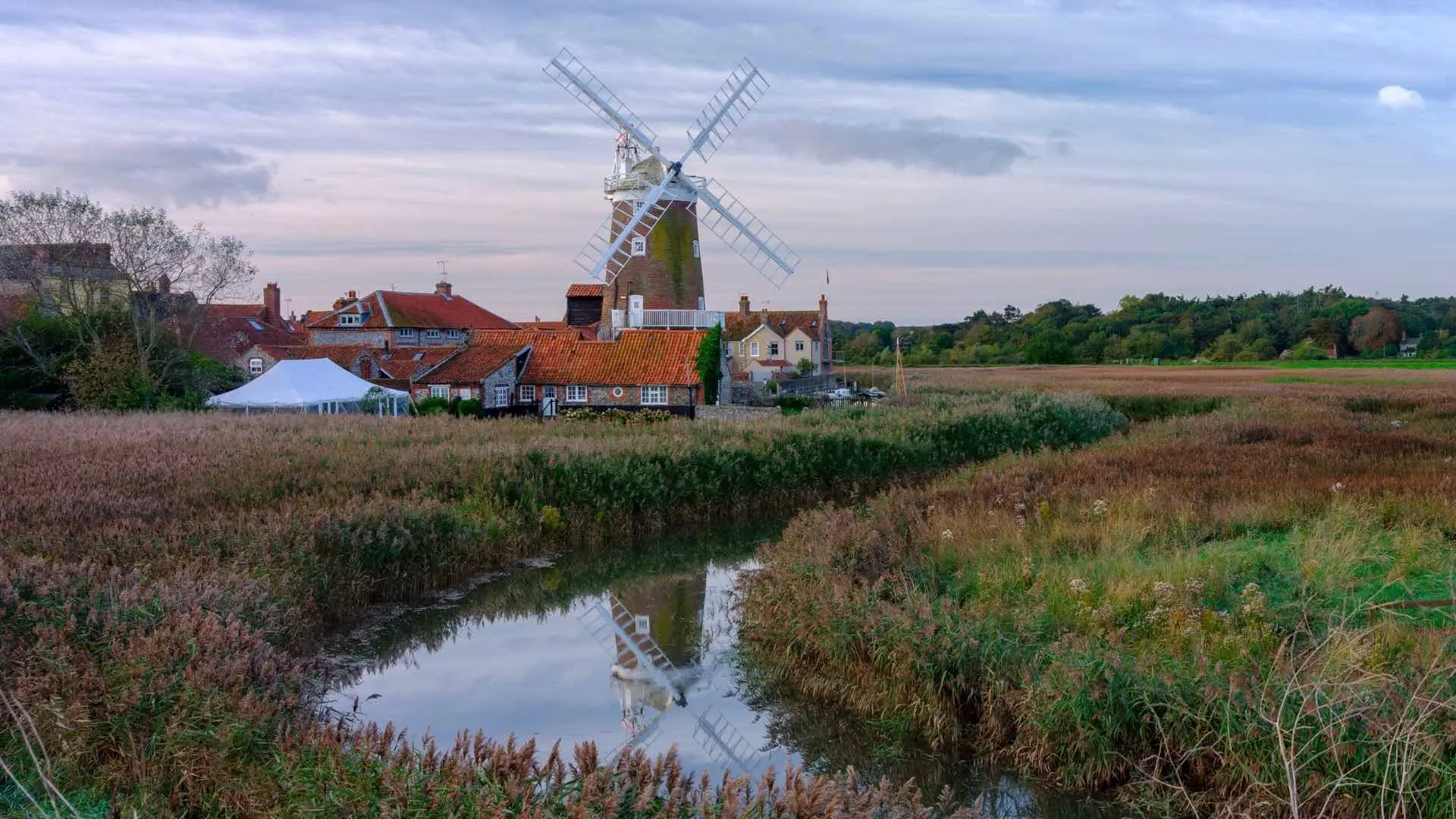 Traditional windmill in the village of Cley-next-the-Sea, Norfolk, surrounded by reed marshes and rustic cottages with red-tiled roofs under a soft, overcast sky.