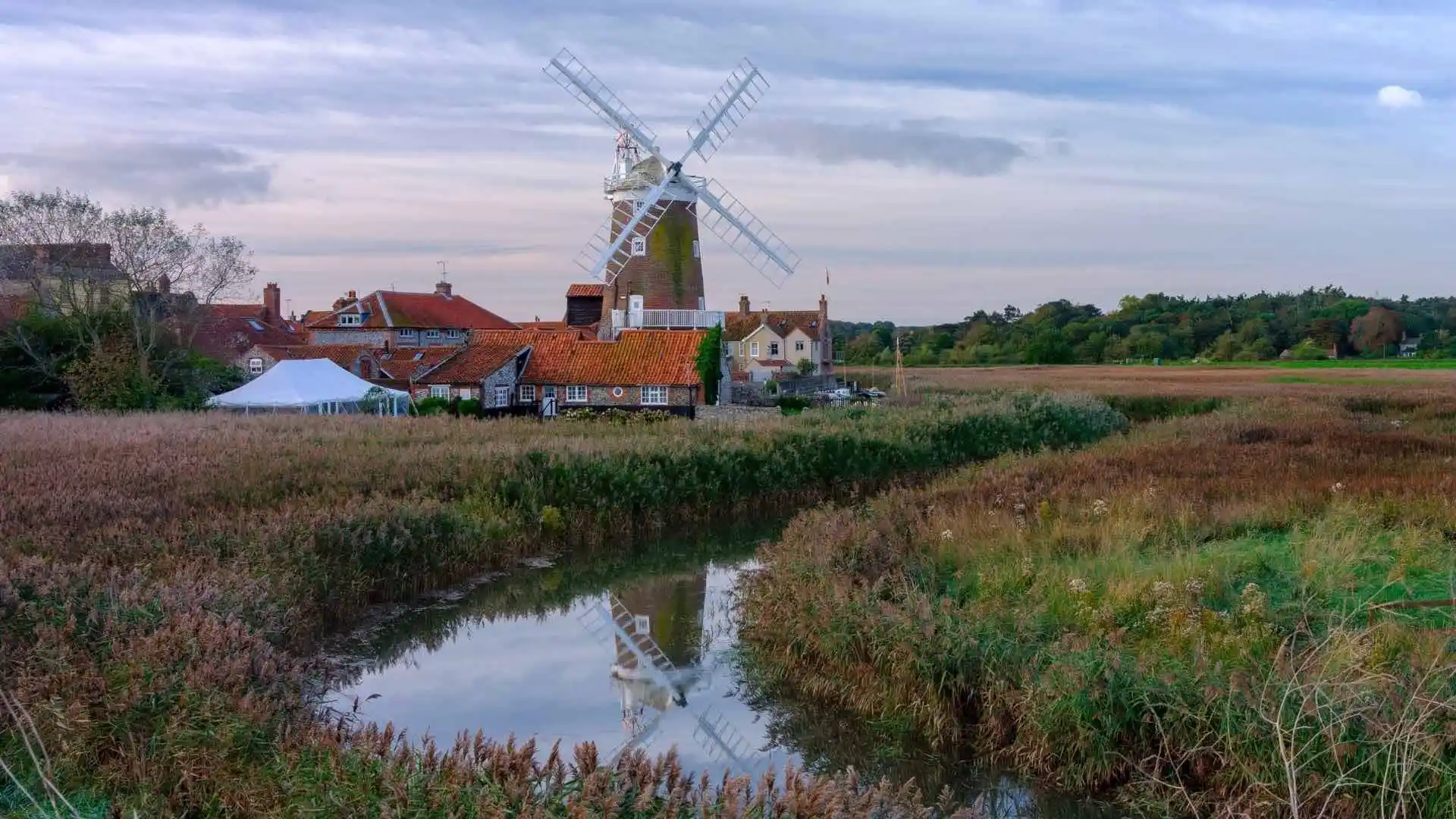 Traditional windmill in the village of Cley-next-the-Sea, Norfolk, surrounded by reed marshes and rustic cottages with red-tiled roofs under a soft, overcast sky.