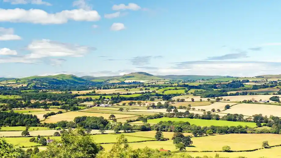 Expansive view of patchwork farmland and rolling green hills under a blue sky with scattered clouds, in the English or Welsh countryside.