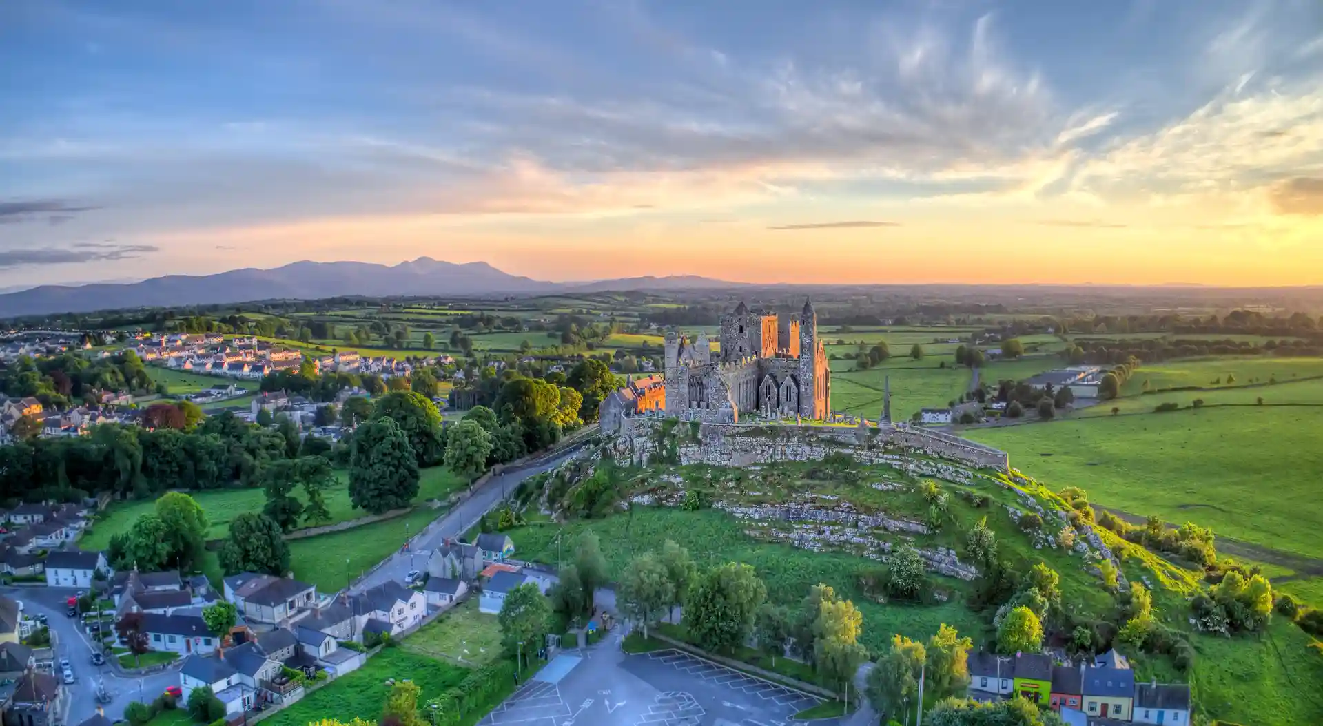 Sunset aerial view of the Rock of Cashel in County Tipperary, Ireland, surrounded by green fields, villages, and distant hills.