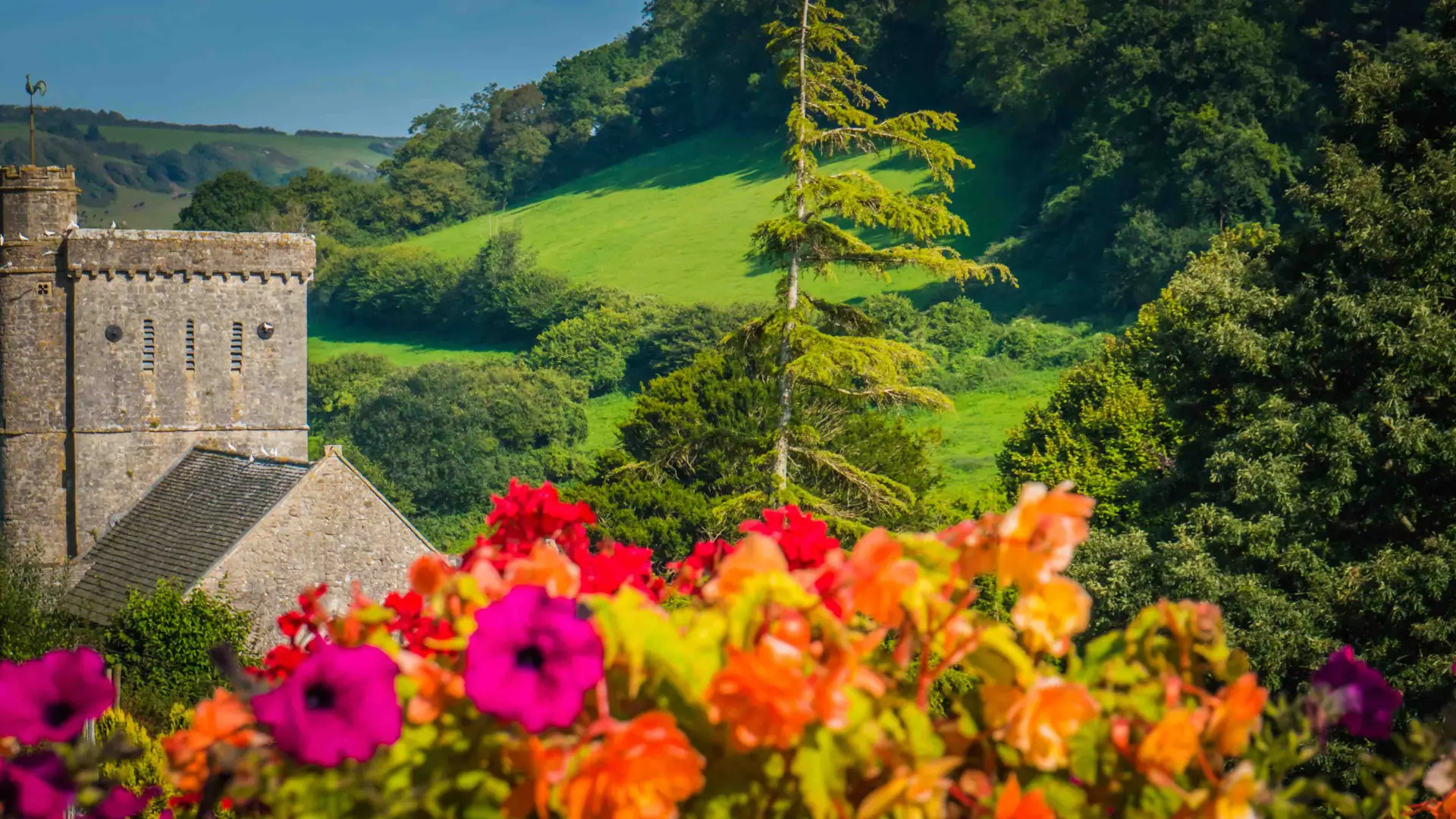 Colourful flowers in the foreground with a historic stone church and lush green countryside in the background on a sunny day.