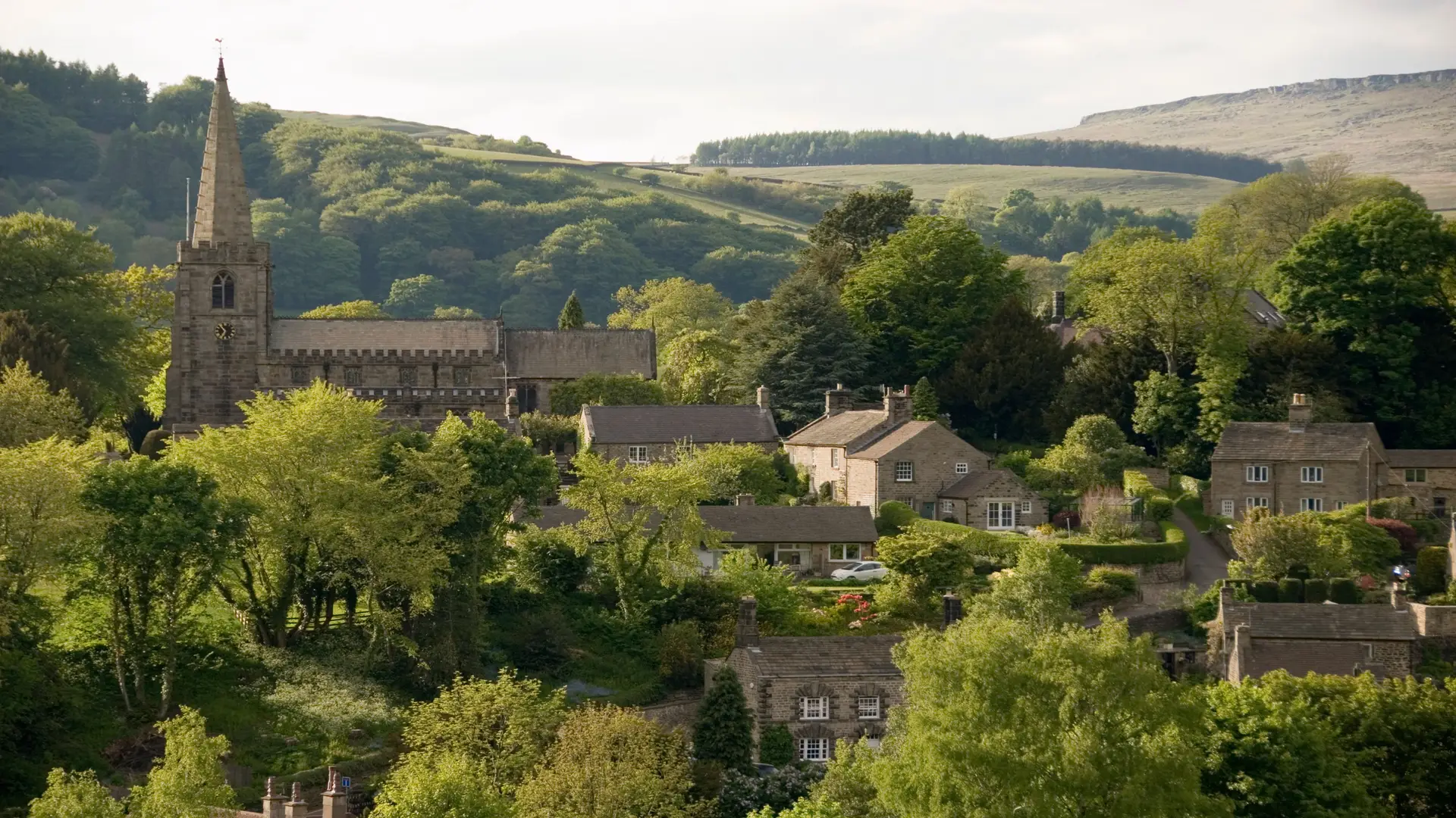 Aerial view of Hathersage village with St Michael and All Angels' Church spire rising amidst stone cottages and autumnal trees, set against the rolling hills of the Peak District in Derbyshire, England.