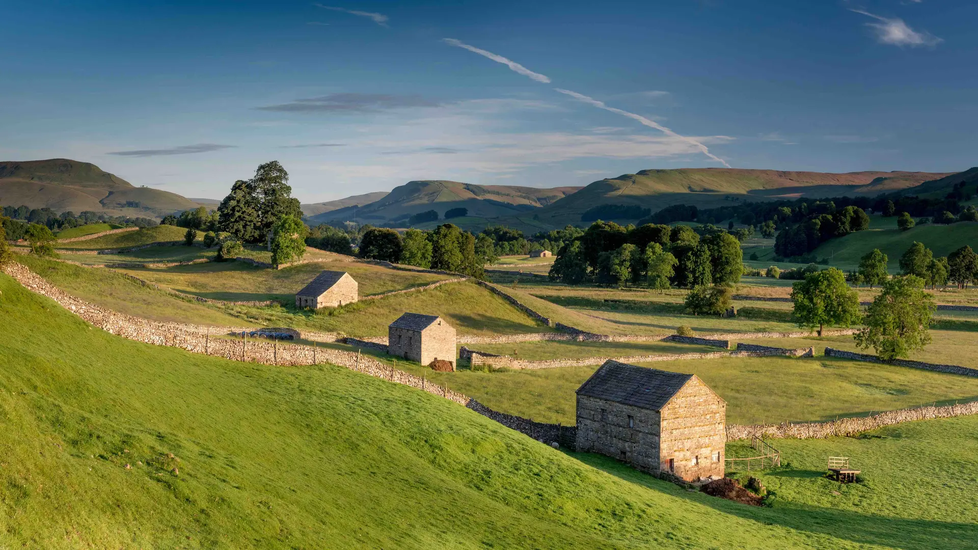 Traditional stone barns and dry-stone walls across rolling green hills in the Yorkshire Dales, with distant moorland rising under a blue sky.