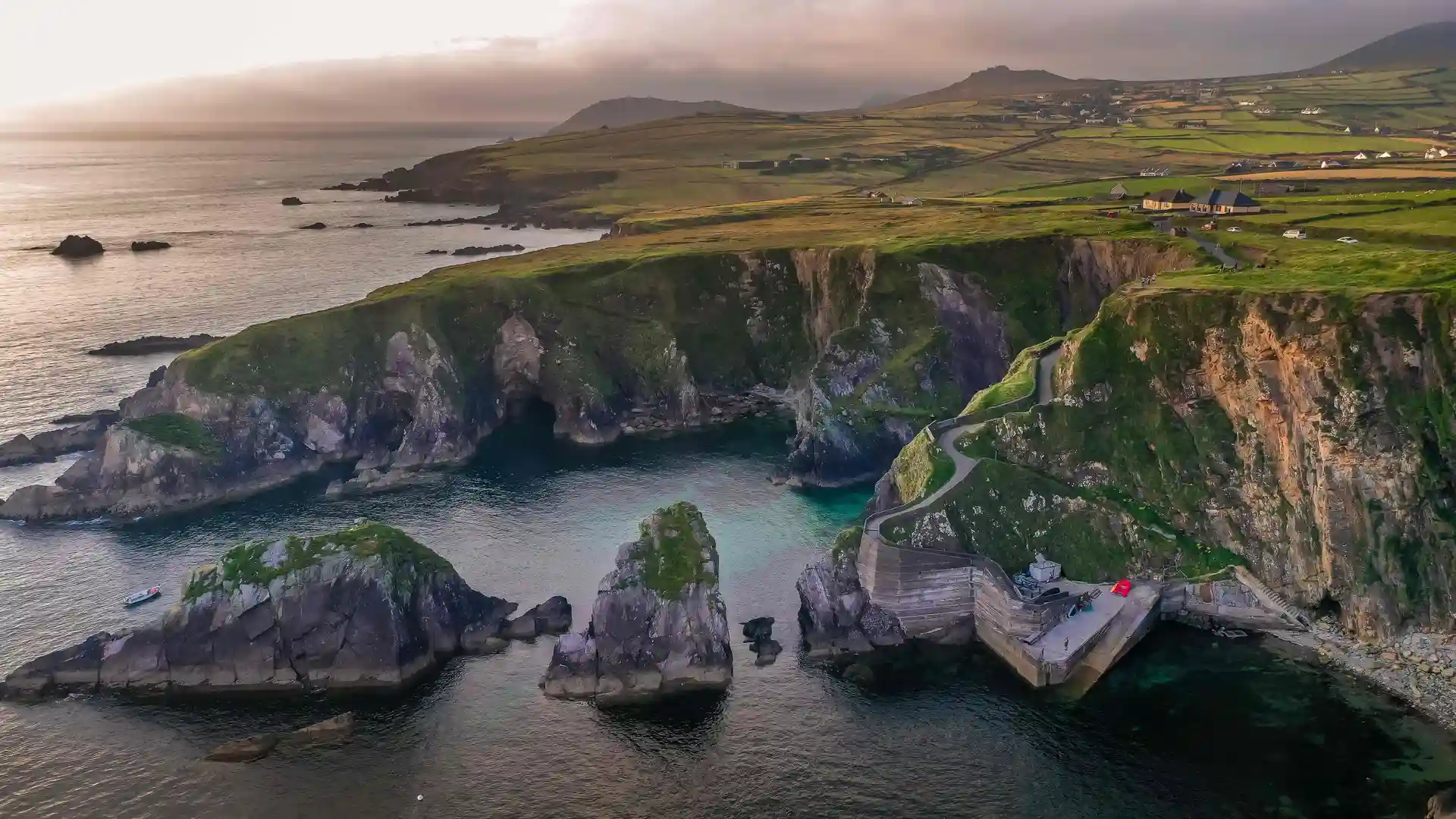Cliffs and harbour on the Dingle Peninsula in the West of Ireland, with Atlantic waters, rocky outcrops, and patchwork farmland under a cloudy sky.
