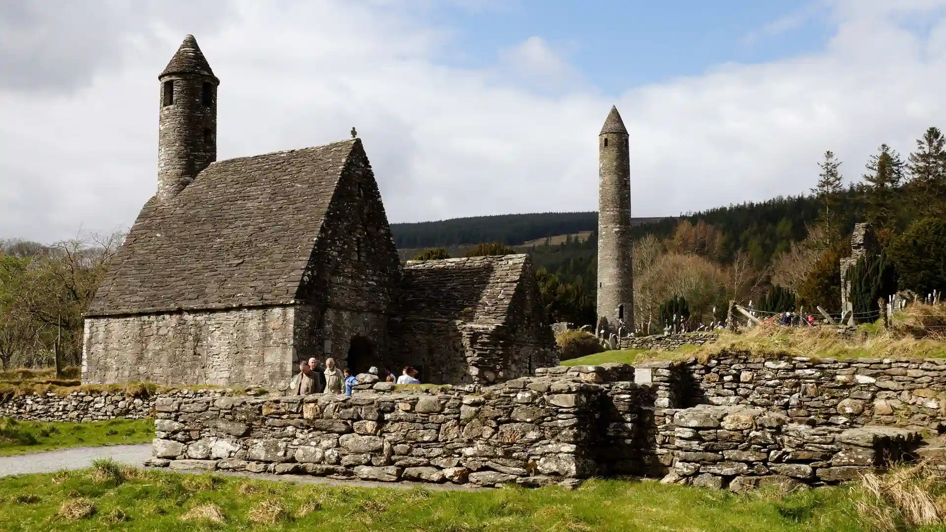 Visitors exploring the monastic site at Glendalough in Ireland’s Ancient East, with a round tower, stone church, and wooded hills in the background.