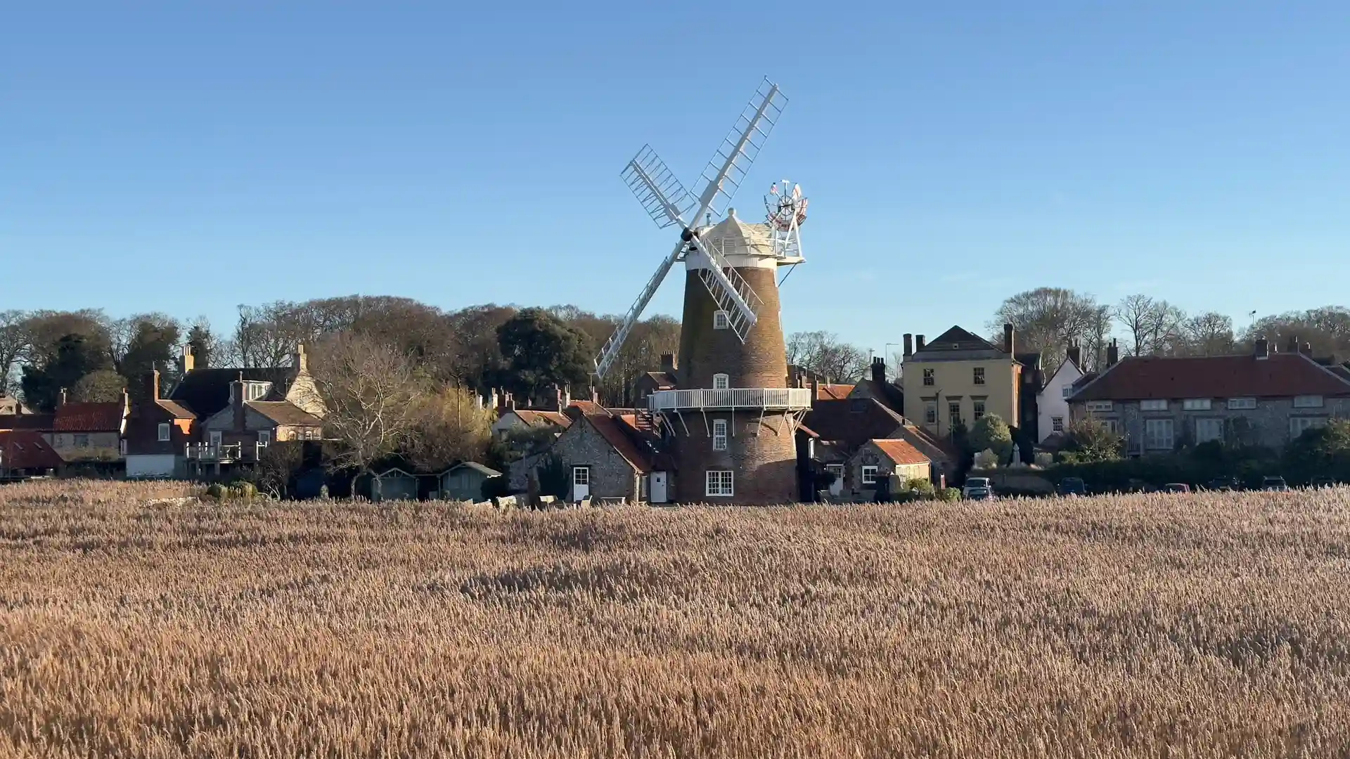 Traditional windmill in the village of Cley-next-the-Sea, Norfolk, surrounded by reed marshes and rustic cottages with red-tiled roofs under a soft, overcast sky.
