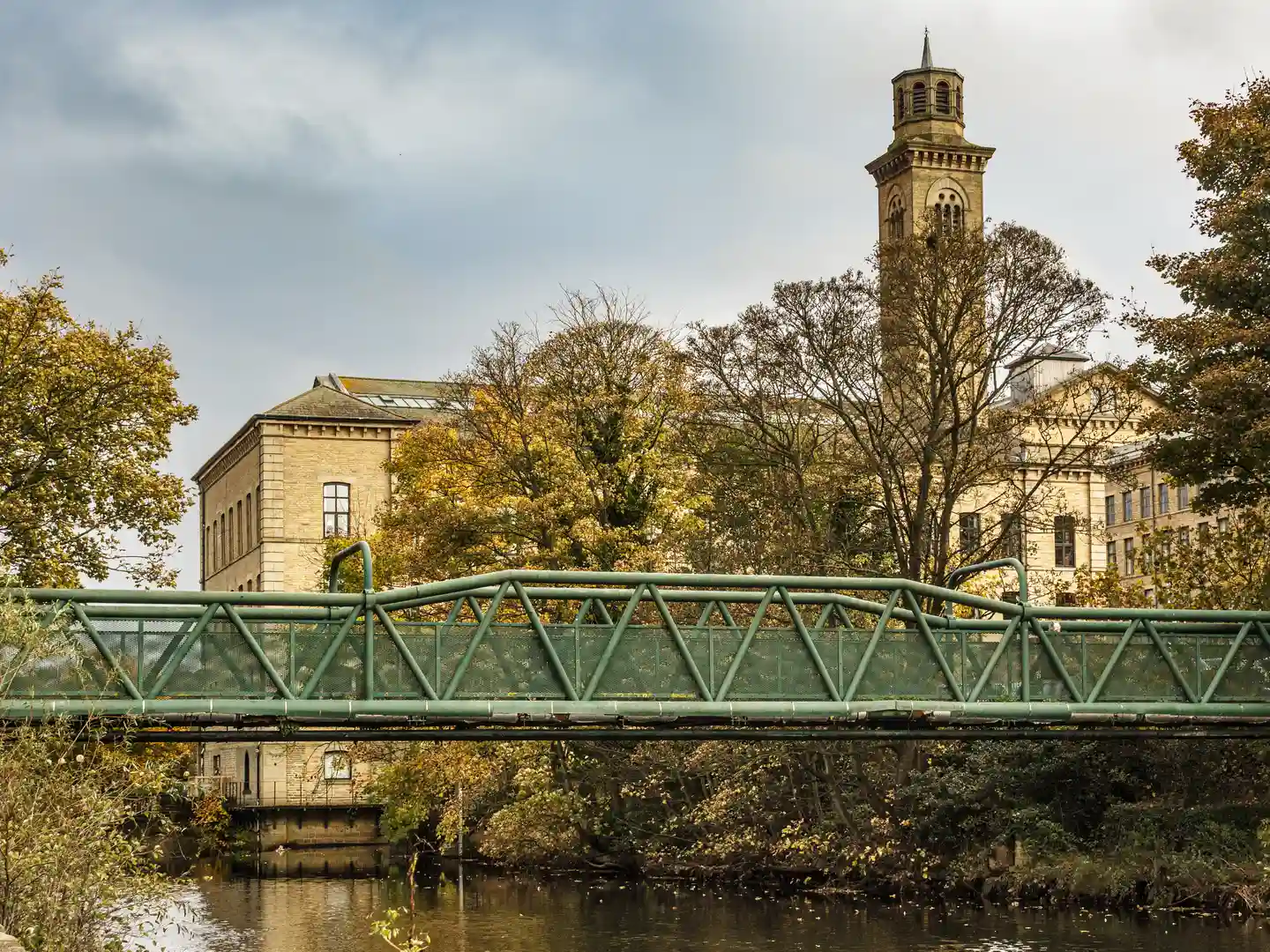 Pedestrian bridge over the River Aire with historic Saltaire mill buildings and tower in autumn light.