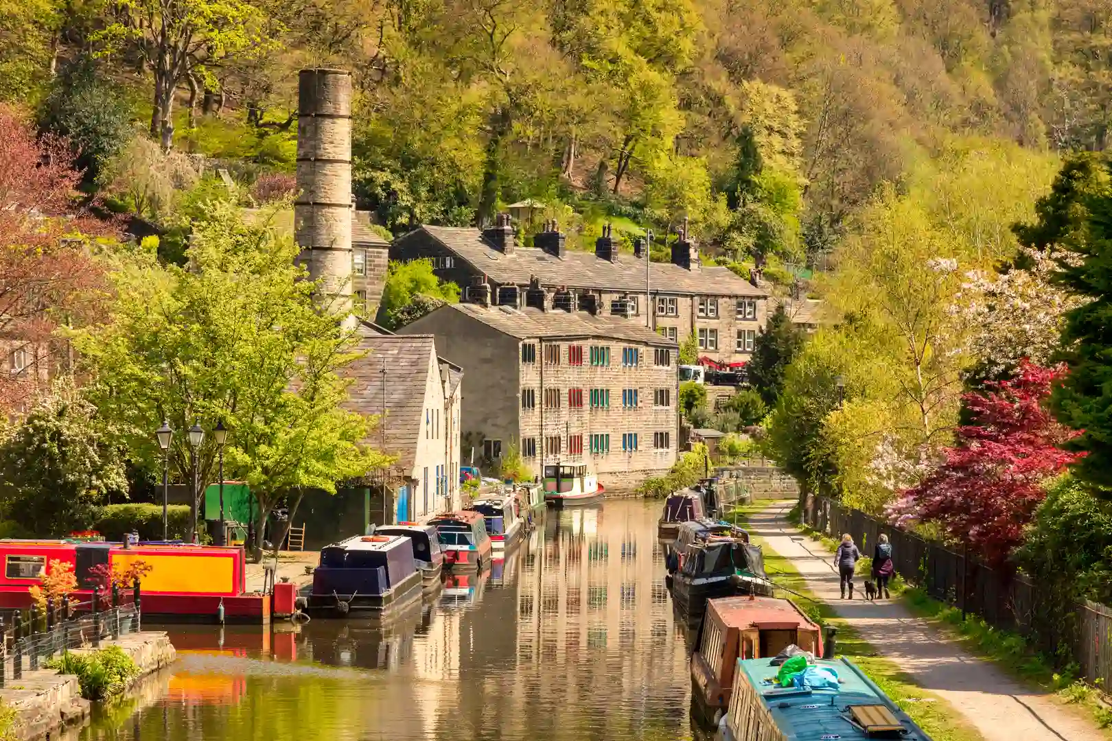 Springtime view of the Leeds and Liverpool Canal at Saltaire in West Yorkshire with narrowboats, stone mills, riverside path and woodland hillside