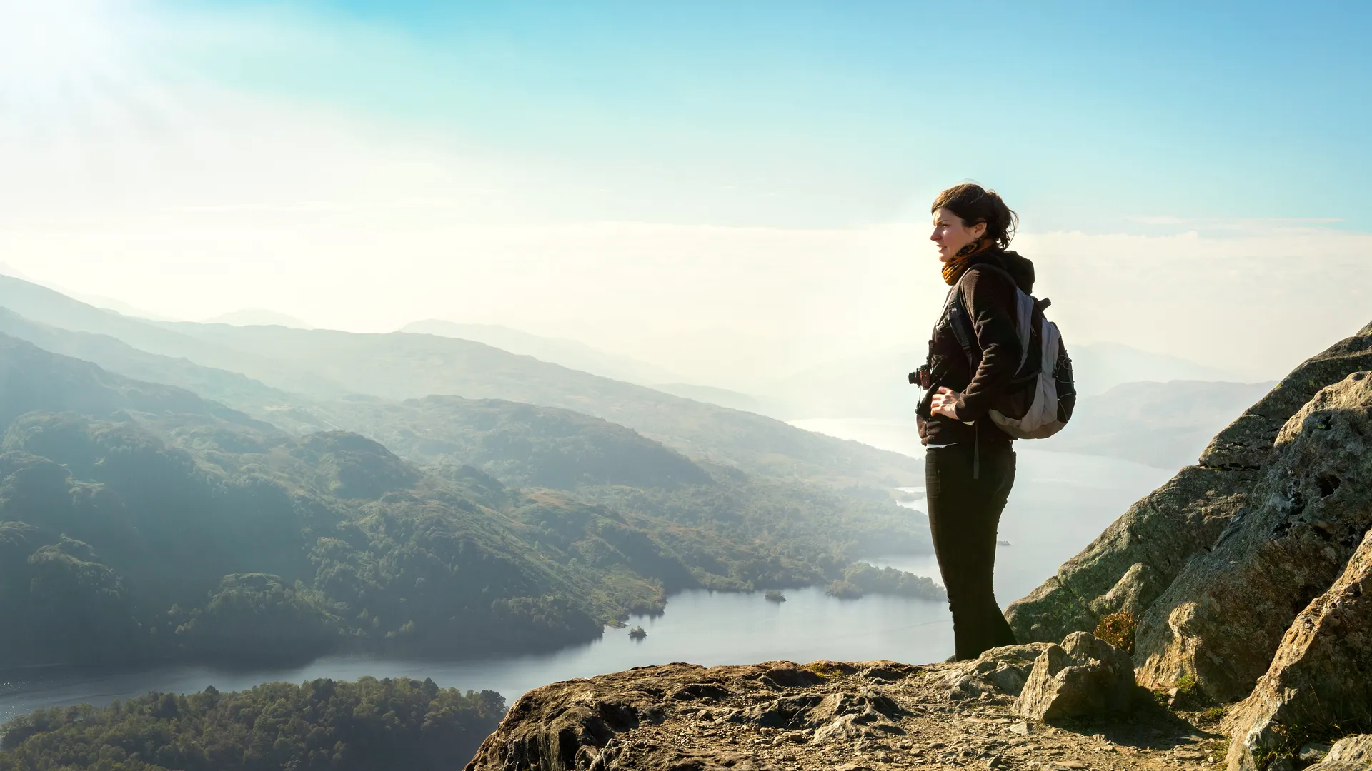 Woman standing on the summit of Ben A’an in Central Scotland, overlooking Loch Katrine and surrounding hills on a clear, hazy morning