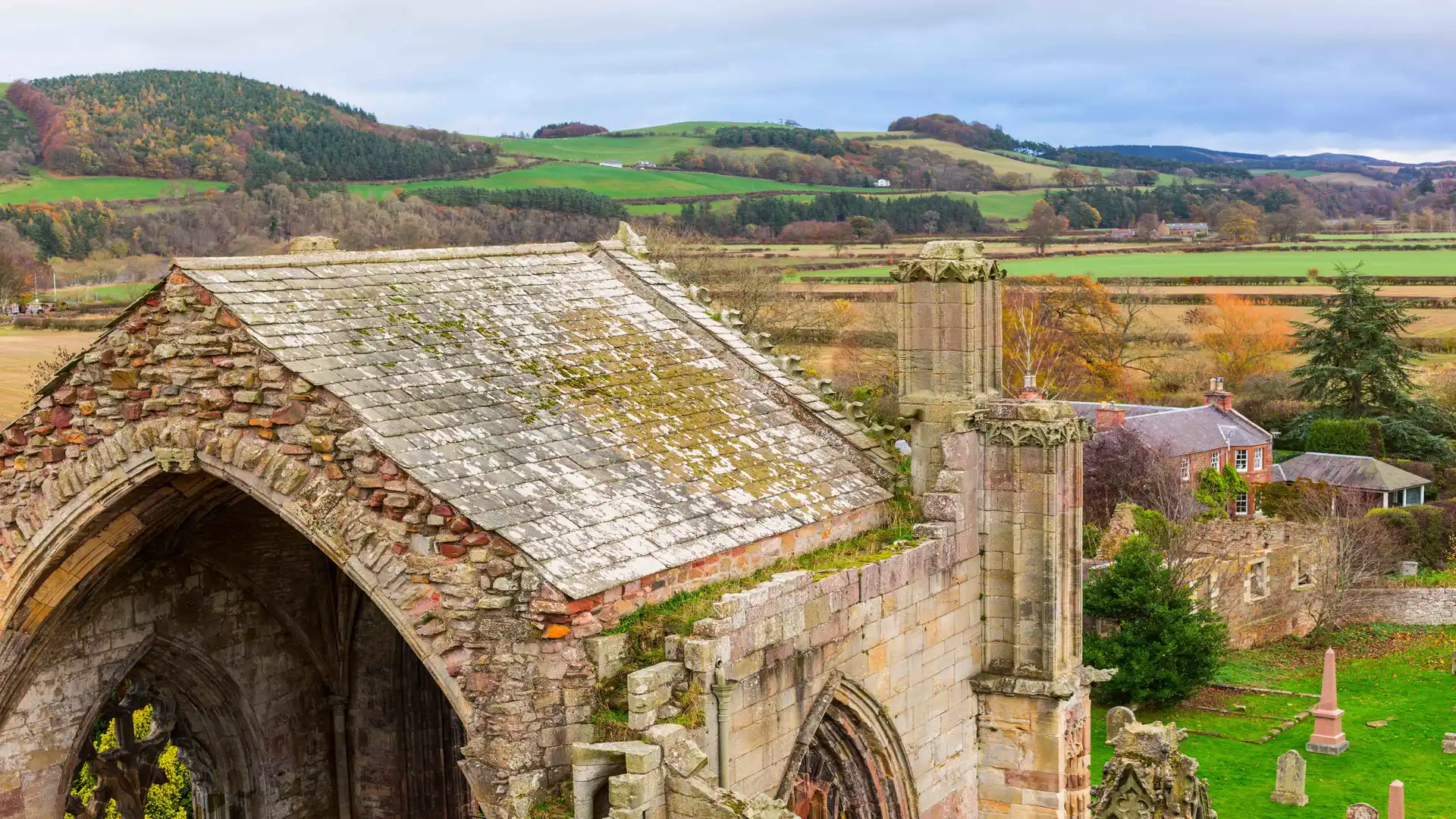 View over Melrose Abbey in the Scottish Borders, with stone ruins, nearby cottages, and rolling farmland under a soft autumn sky.