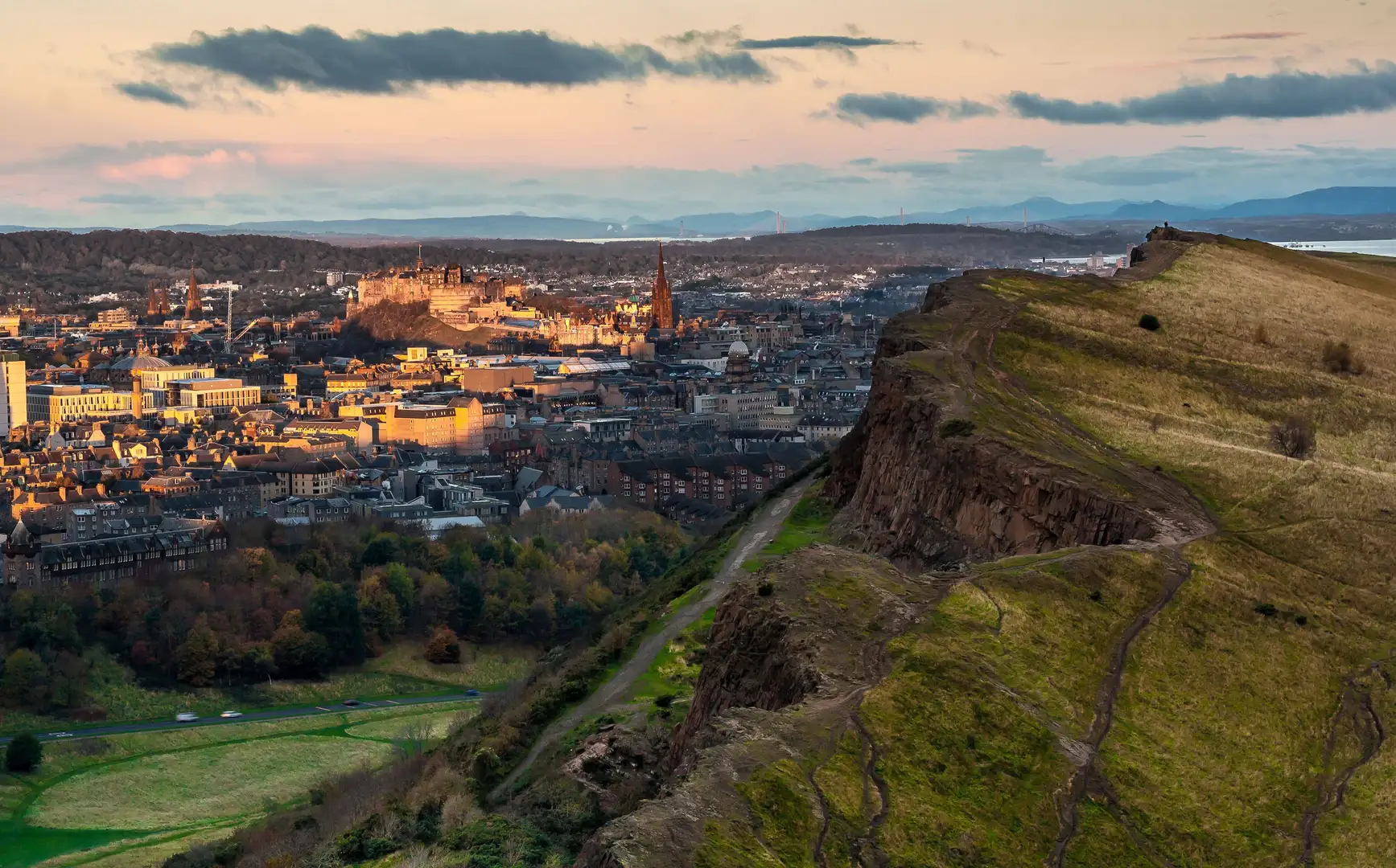 View from Salisbury Crags in Holyrood Park, Edinburgh, with walking trails in the foreground and the city skyline, including Edinburgh Castle, at sunset.