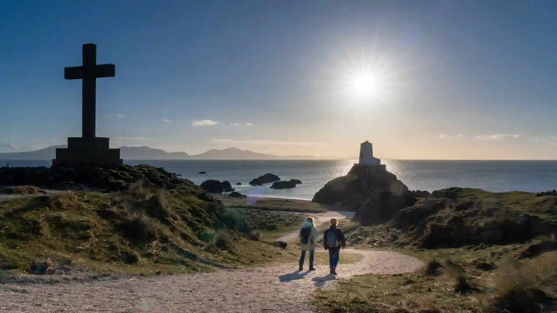 Walkers approaching Tŵr Mawr lighthouse on Llanddwyn Island, Isle of Anglesey, with Celtic cross, coastal views, and golden evening light.