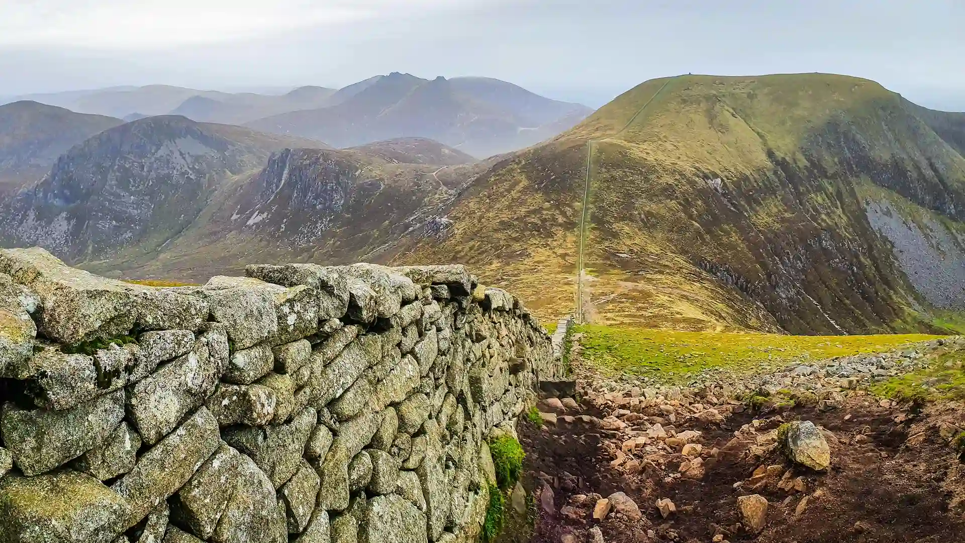 A rugged mountain path follows the historic Mourne Wall as it winds through granite peaks and rolling uplands in the Mourne Mountains, Northern Ireland, under a hazy sky.