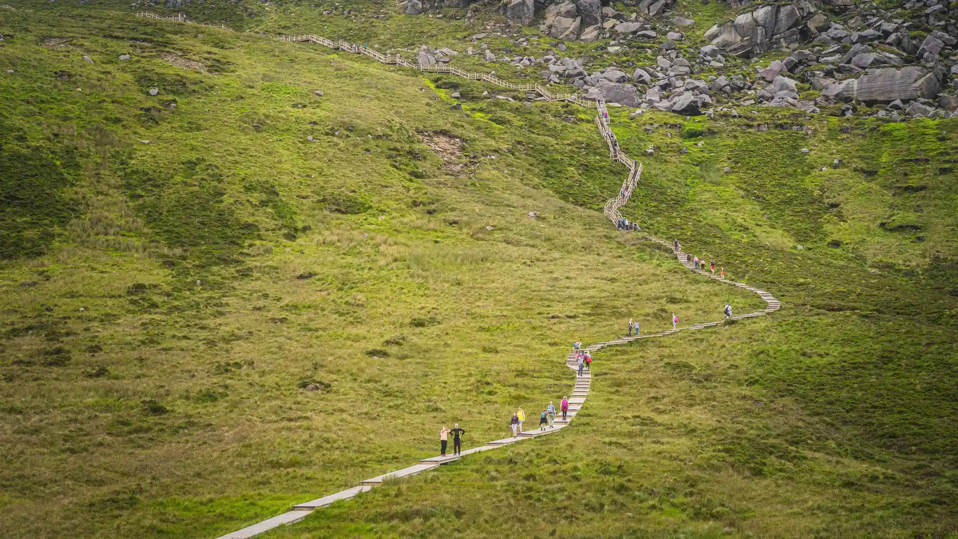 Walkers ascending the wooden boardwalk trail on Cuilcagh Mountain, known as the “Stairway to Heaven,” surrounded by rolling green hills and scattered rocks in the Fermanagh Lakelands of Northern Ireland.