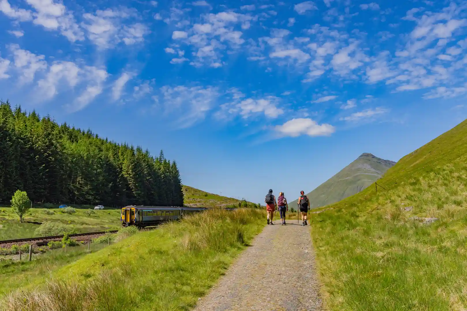 Three hikers walk along the West Highland Way in Scotland on a sunny day, passing a train line and green hills near Tyndrum beneath a bright blue sky.