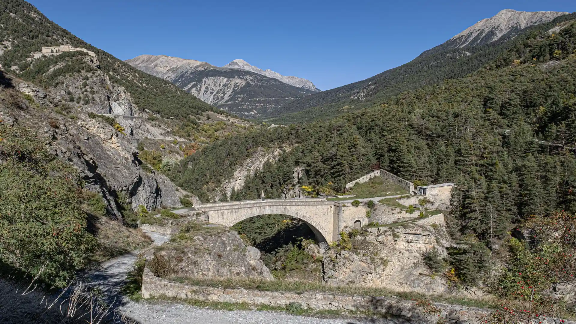 Stone Vauban-era bridge crossing a mountain gorge in the Upper Durance Valley near Briançon, surrounded by forested slopes and high peaks in the French Alps.