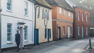 A quiet street in Nayland, Suffolk, lined with colourful historic buildings and Georgian windows, within the Dedham Vale National Landscape. A walker passes along the pavement on a calm morning.