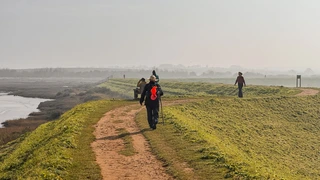 Walkers on a raised coastal path beside saltmarsh and tidal water on the Norfolk Coast Path, with soft winter light and misty horizons.