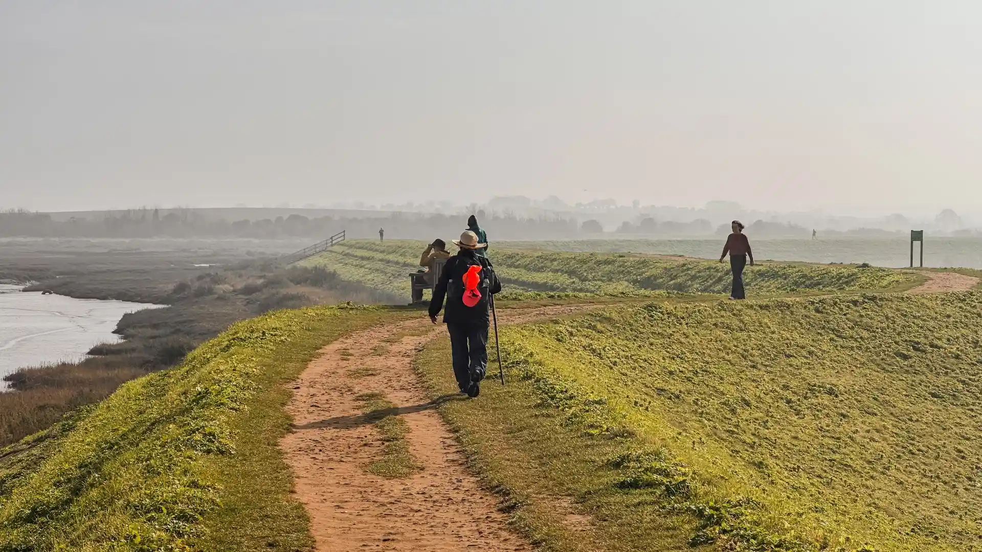 Walkers on a raised coastal path beside saltmarsh and tidal water on the Norfolk Coast Path, with soft winter light and misty horizons.