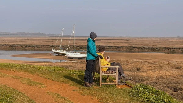 Walkers resting beside saltmarsh along the Norfolk Coast Path, with boats moored on a tidal creek.