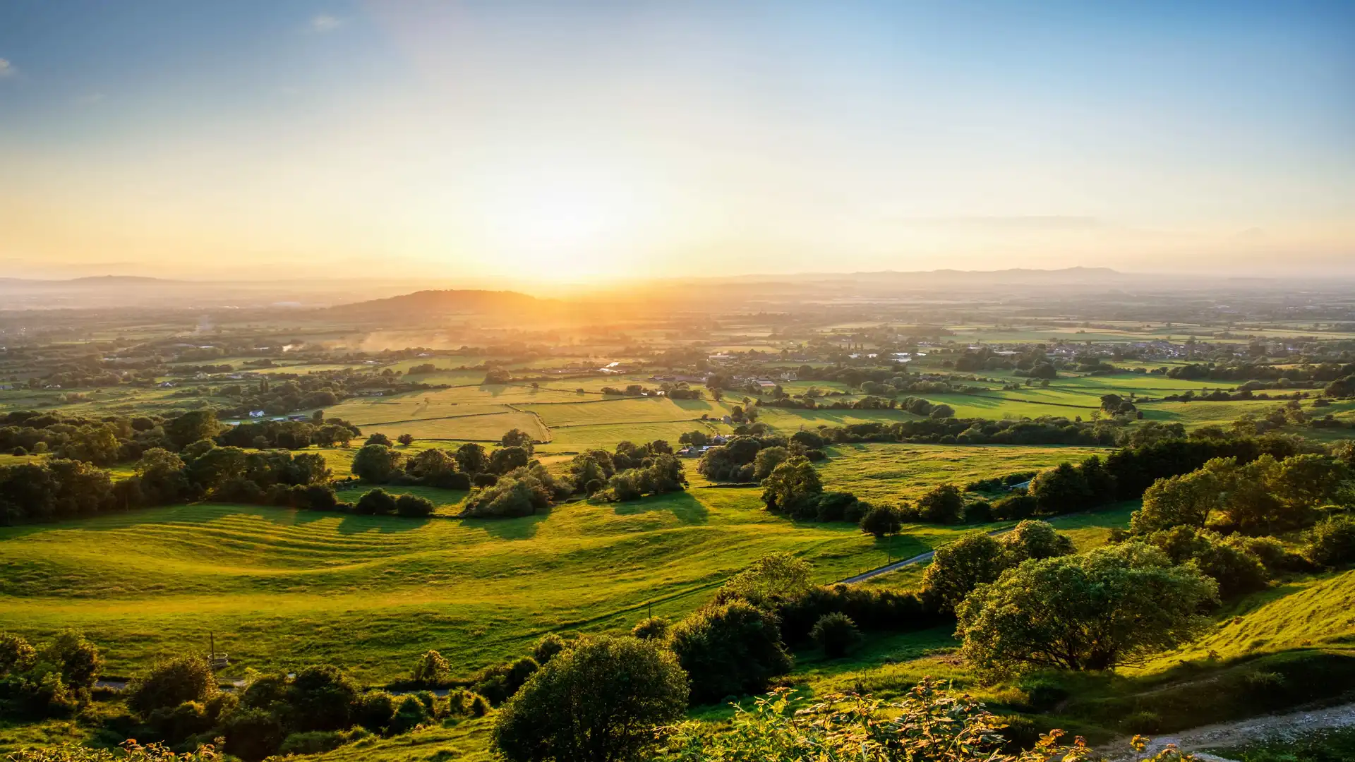 Rolling hills and patchwork farmland at sunset in the Cotswolds National Landscape.