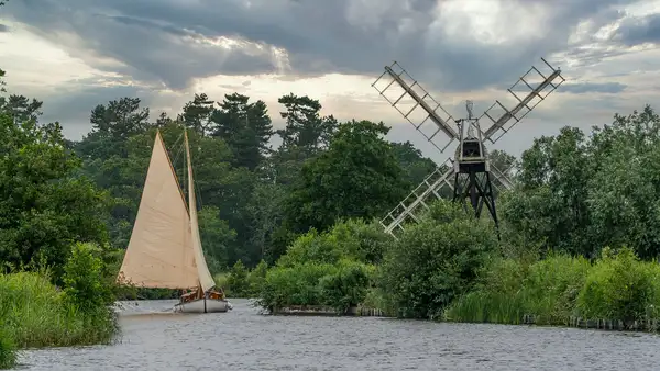 Traditional sailing boat passing a riverside windmill on a tree-lined waterway in The Broads National Park, Norfolk.