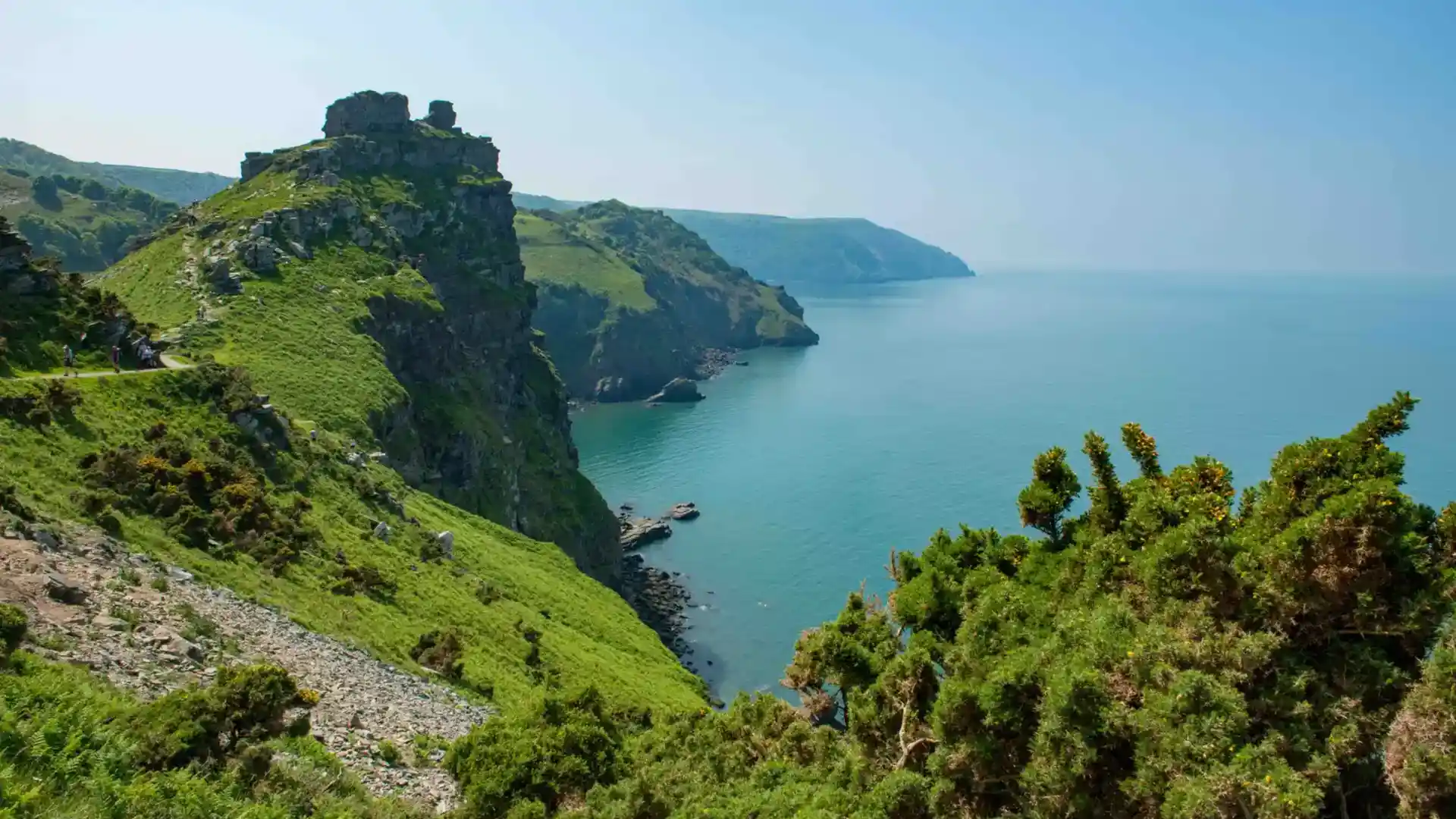 Rugged clifftop walking path above the Atlantic on the North Devon coast, with steep green cliffs, headlands, and calm blue sea below along the South West Coast Path.