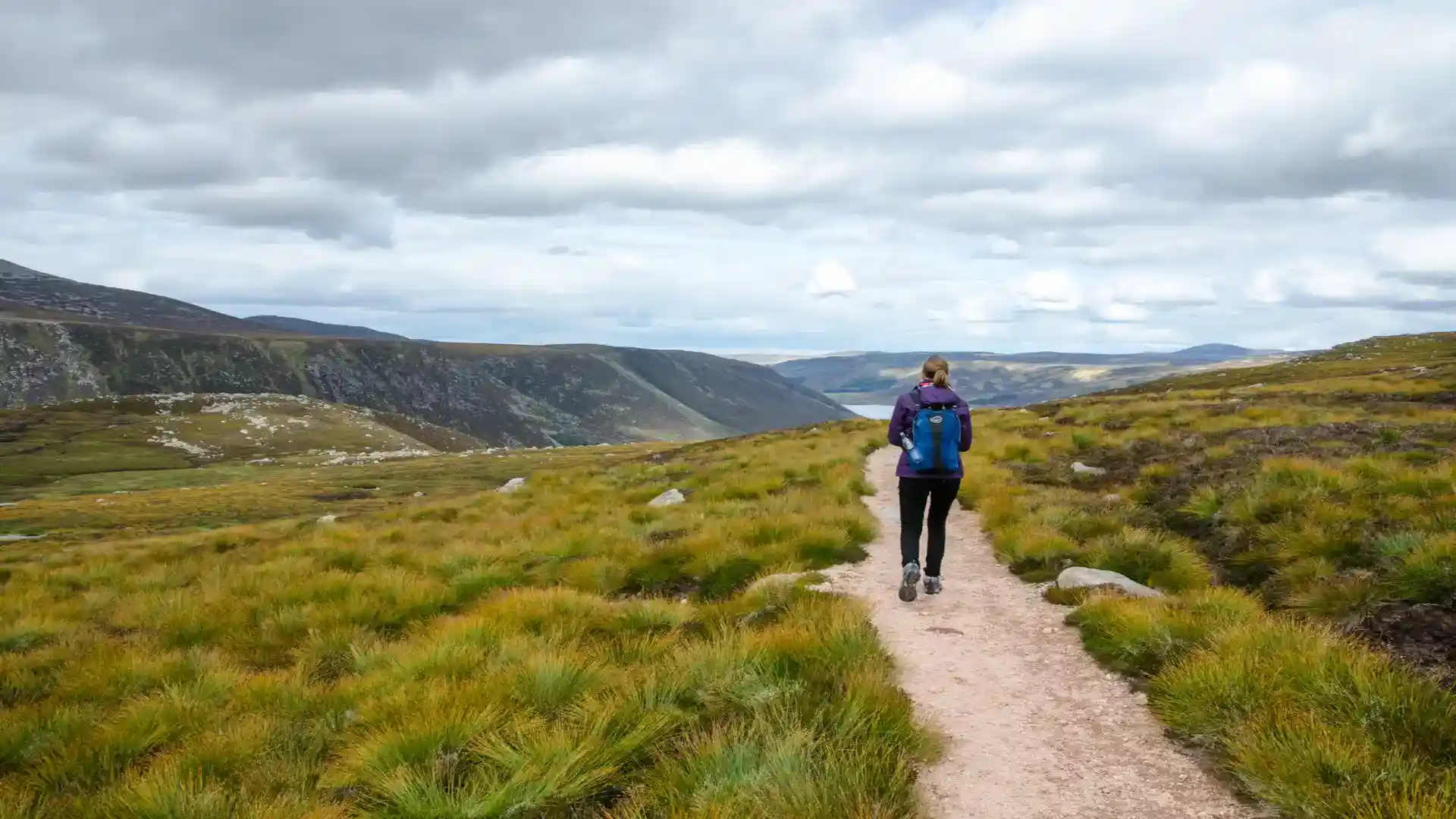 A hiker walks along a mountain trail in Cairngorms National Park, Scotland, surrounded by heather-covered hills and sweeping glen views under a cloudy sky.