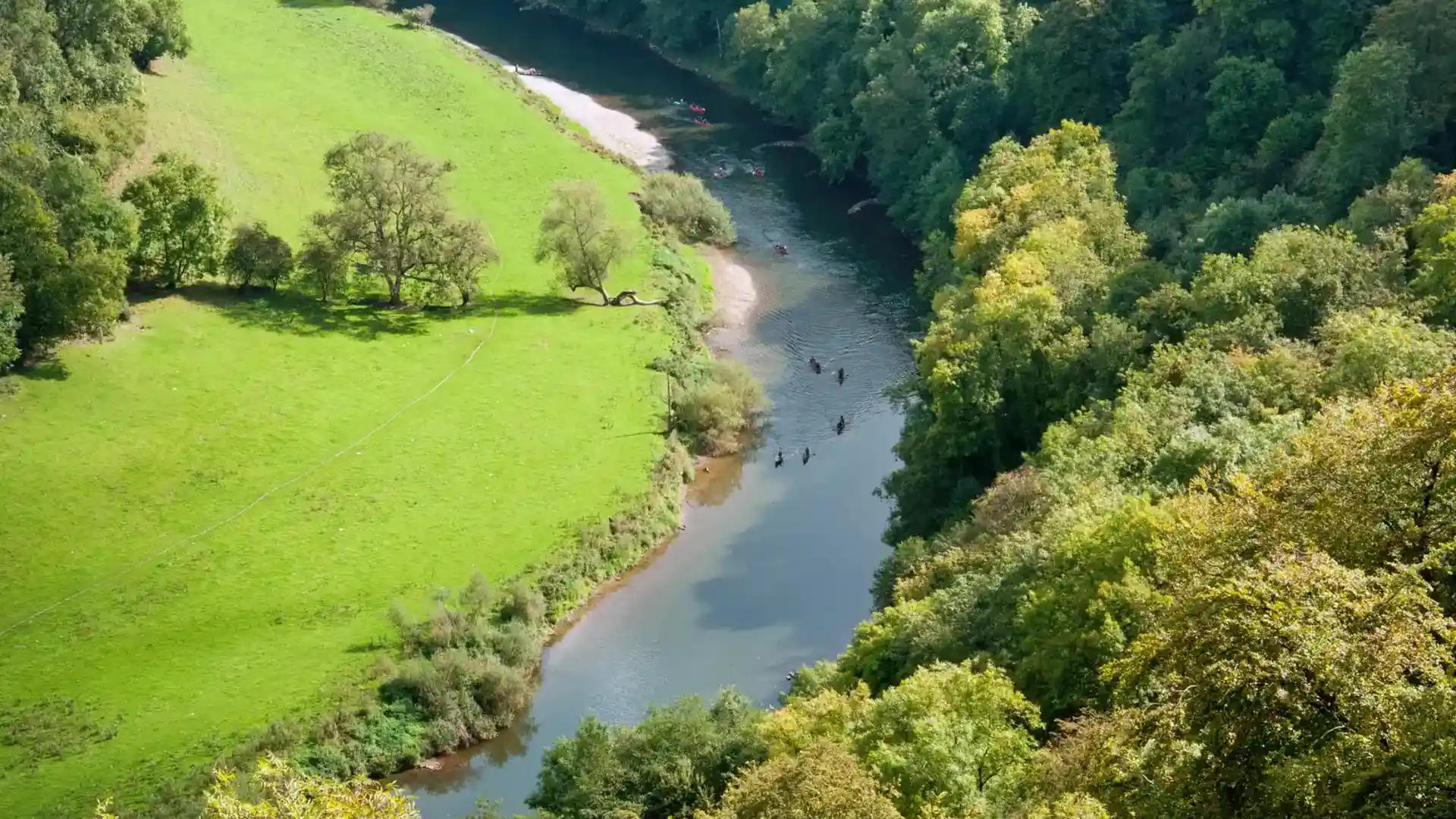 Canoeists paddling along a winding stretch of the River Wye, surrounded by green meadows and dense woodland in the Wye Valley National Landscape.