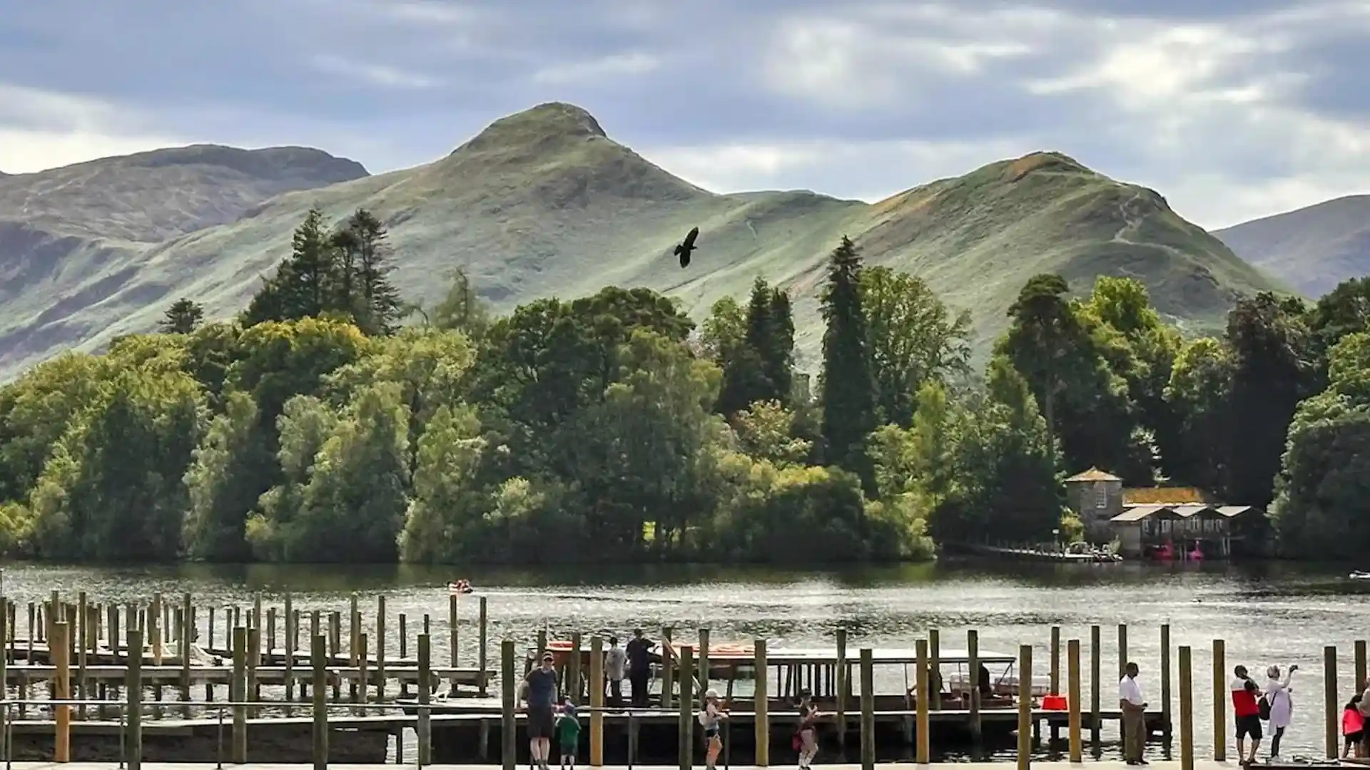 View across Derwentwater towards Cat Bells, with a lakeside jetty in the foreground, calm water, woodland, and the green fells of the Lake District under a soft, cloudy sky.