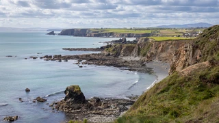 Cliffs and rocky shoreline along the Copper Coast in County Waterford, Ireland, with calm sea and patchwork farmland above the coastal edge.