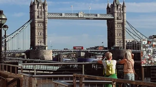 Two people standing by the River Thames looking toward Tower Bridge with a red London bus crossing.