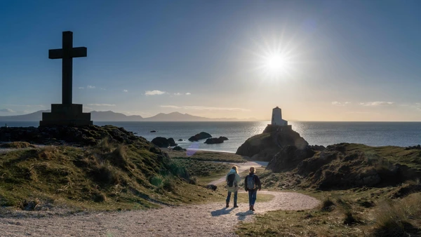Walkers descending towards Llanddwyn Island lighthouse on the Anglesey Coast Path with Snowdonia mountains on the horizon.