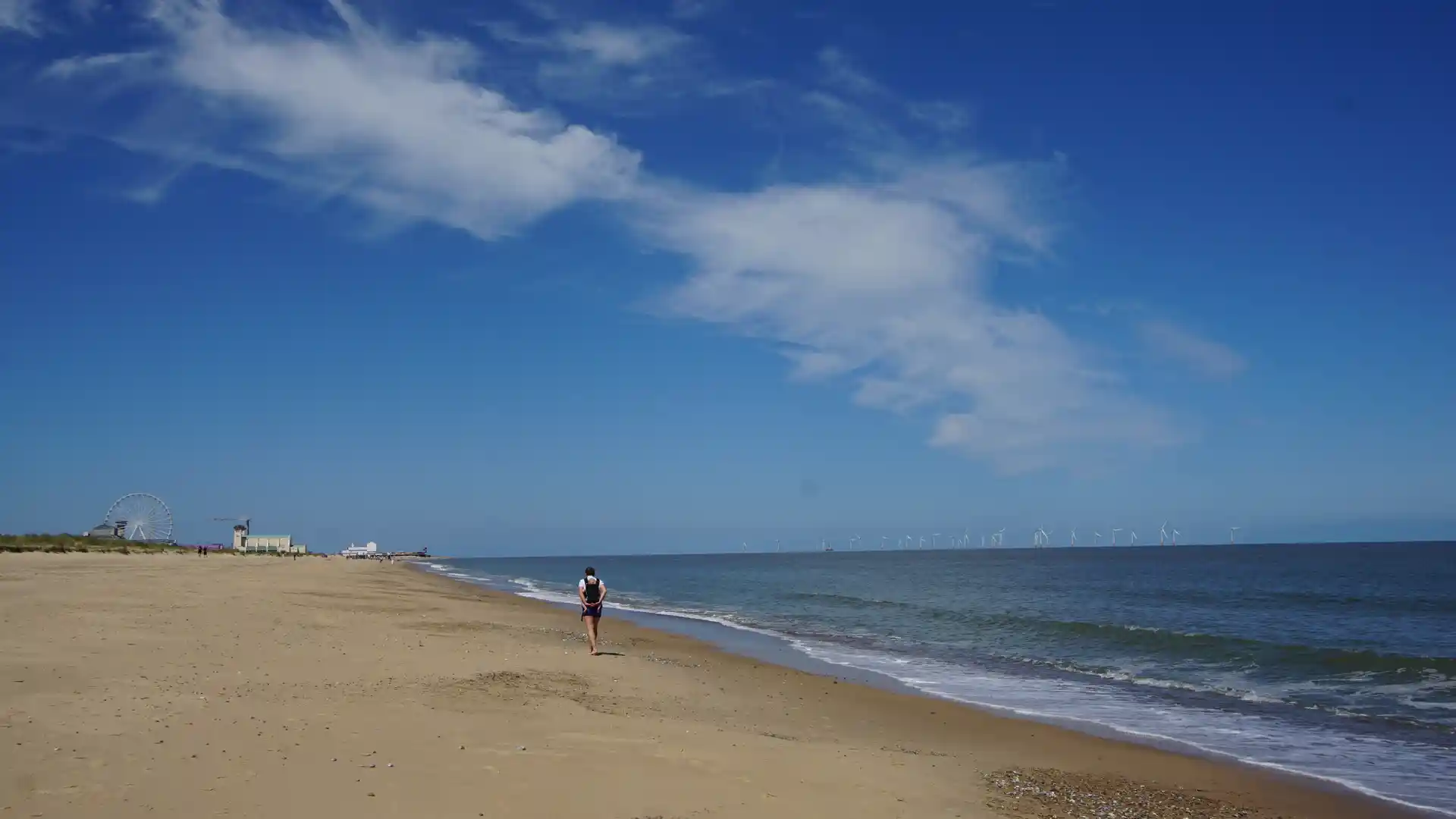 Walker on Great Yarmouth beach along the Norfolk Coast Path with sea, wide sky, and distant wind turbines.