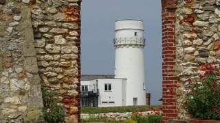 Old Hunstanton Lighthouse viewed through a stone arch on the Norfolk Coast Path with coastal buildings and flowers in the foreground.