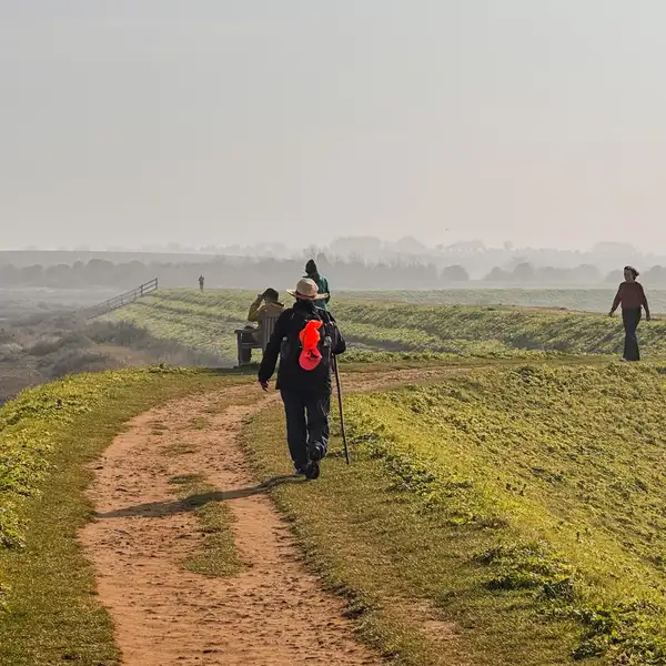Walkers on a raised coastal path beside saltmarsh and tidal water on the Norfolk Coast Path, with soft winter light and misty horizons.