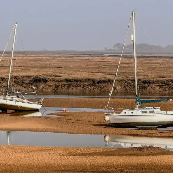 Two sailboats resting on tidal sandbanks at low tide on the Norfolk Coast, surrounded by wide saltmarsh and calm shallow water, with soft coastal light and a hazy horizon.