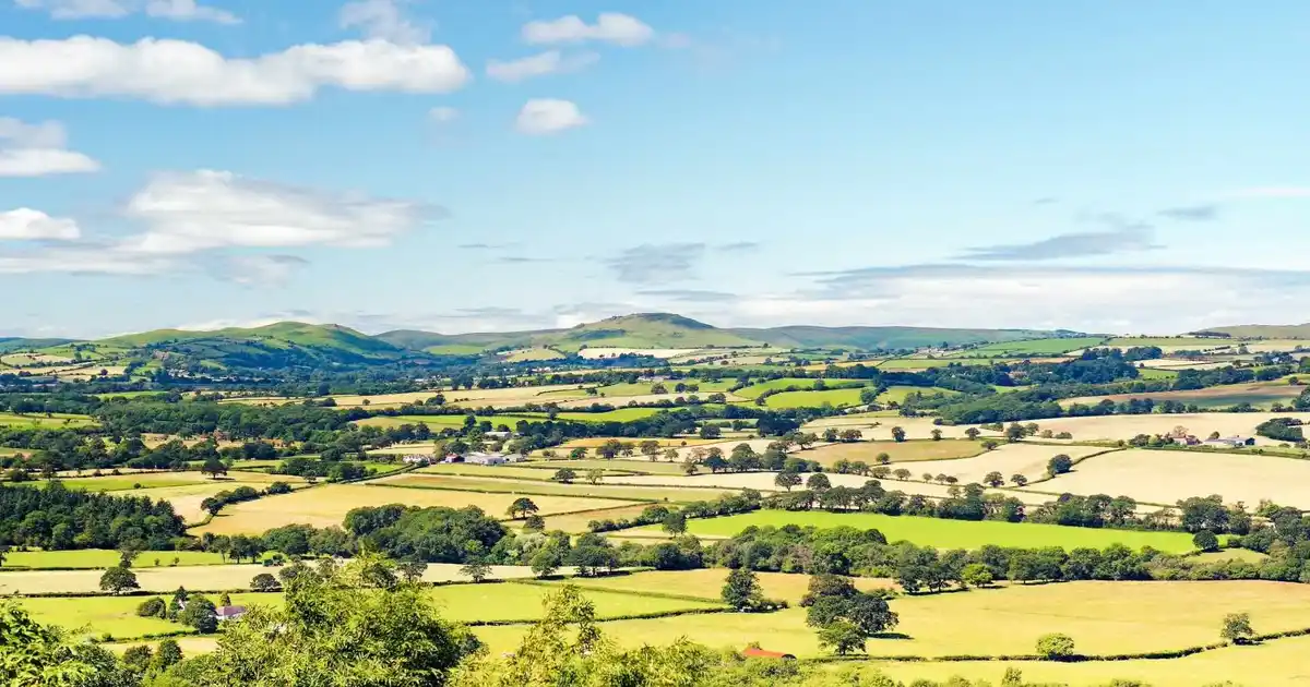 Expansive view of patchwork farmland and rolling green hills under a blue sky with scattered clouds, in the English countryside.
