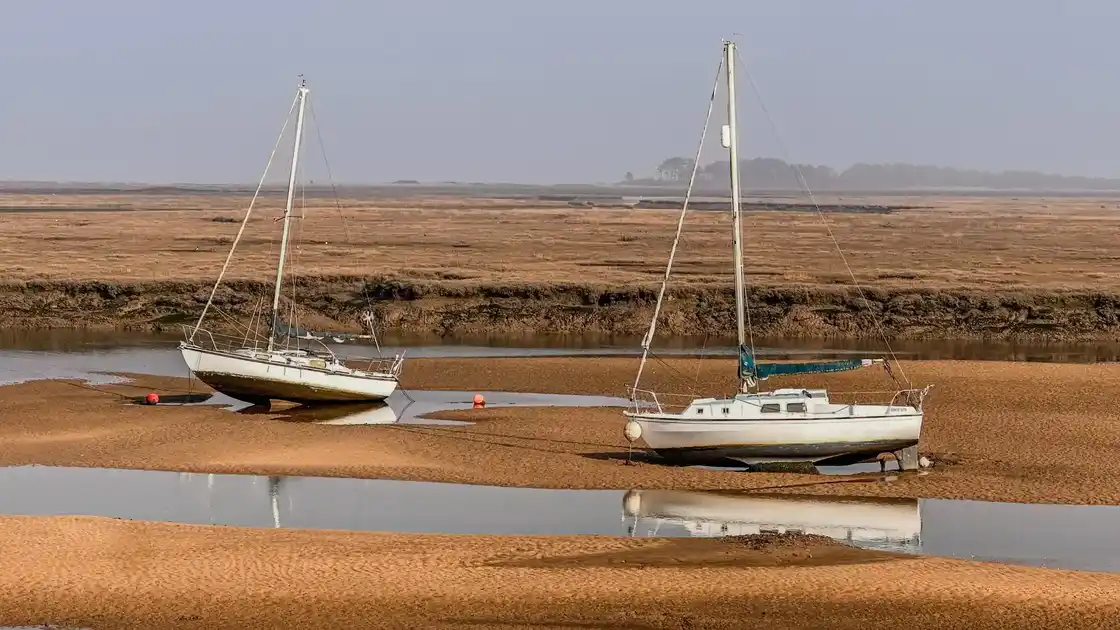 Two sailboats resting on tidal sandbanks at low tide on the Norfolk Coast, surrounded by wide saltmarsh and calm shallow water, with soft coastal light and a hazy horizon.
