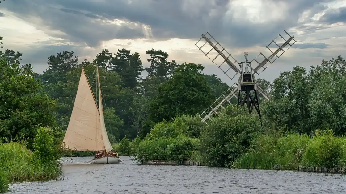 Traditional sailing boat passing a riverside windmill on a tree-lined waterway in The Broads National Park, Norfolk.