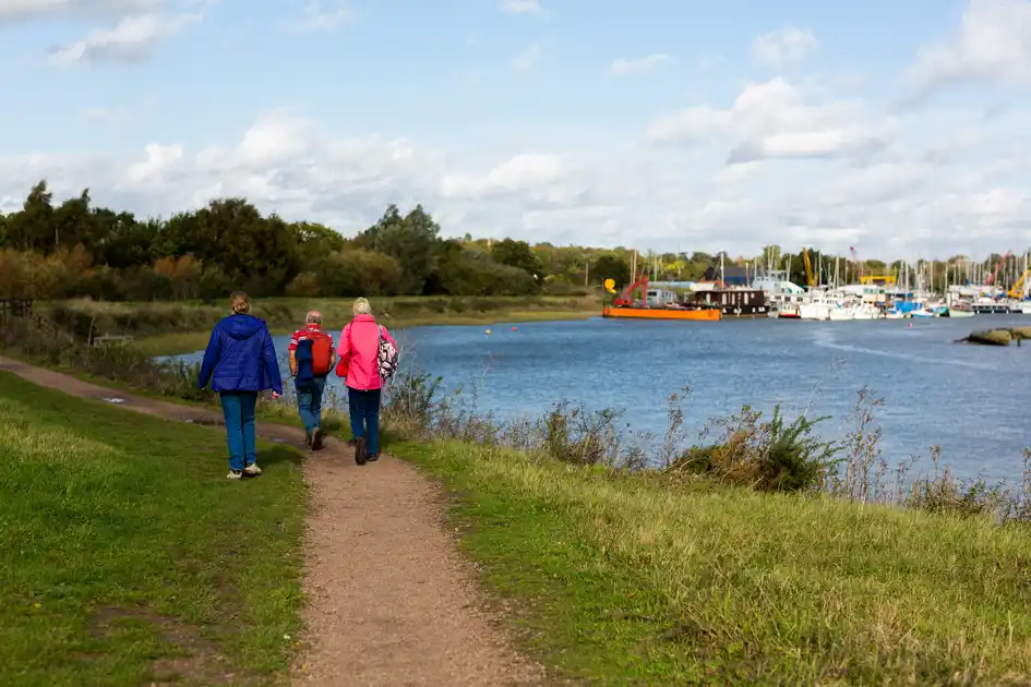 Walkers following a riverside path beside boats and open countryside in East Anglia.